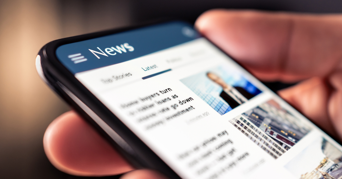 Hand holding a mobile phone. The screen shows news. None of the news stories are legible.